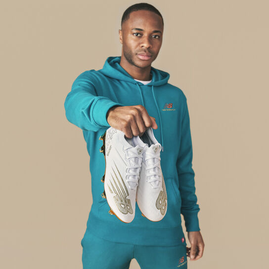 Joueur de foot dans un haut à capuche bleu sarcelle tenant des chaussures à crampons blanches
