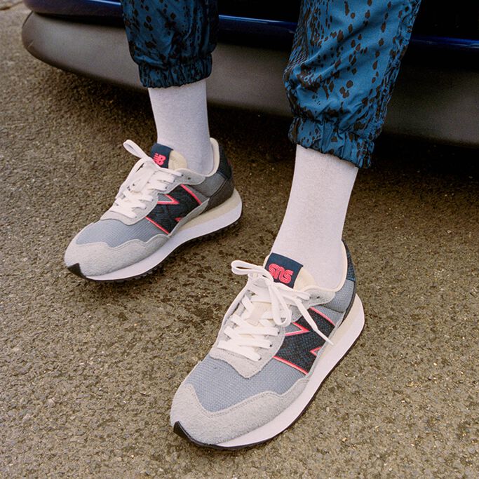 Une image agrandie à partir des genoux d'un homme appuyé sur une voiture et qui porte des chaussures de sport 'Blue Racer' 237 NB x SNS grises et bleu marine rehaussées d'accents magenta