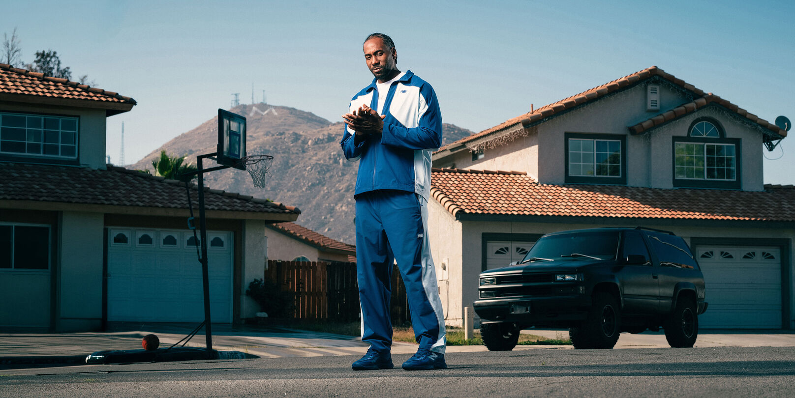 Une personne v&ecirc;tue d&rsquo;un ensemble de surv&ecirc;tement bleu et blanc et de chaussures de sport assorties se tient dans une rue de banlieue, avec des maisons et des montagnes en arri&egrave;re-plan.