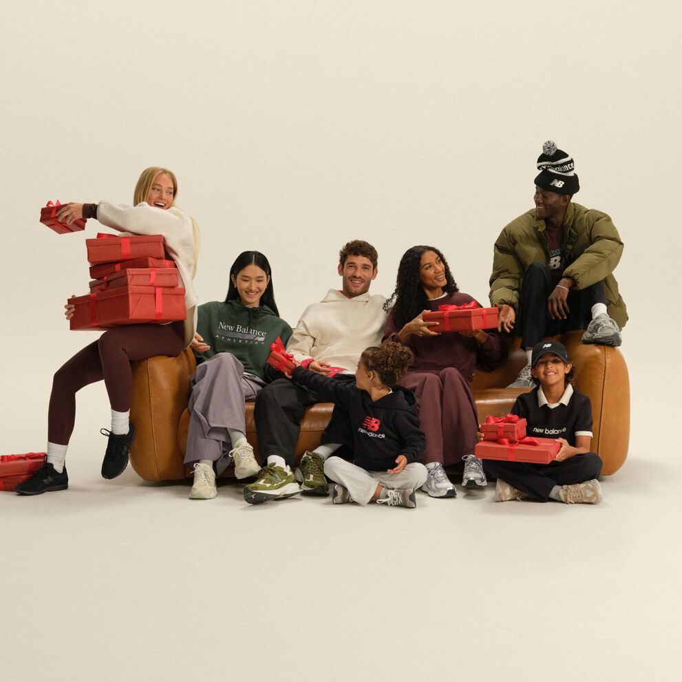 Group of adults and children in casual athletic apparel gathered around a sofa, exchanging red gift boxes in a festive studio setting.