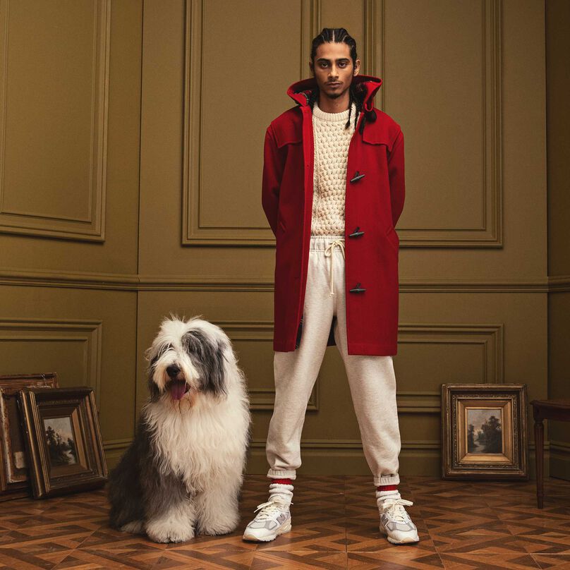 Un homme debout dans un bel environnement intérieur portant des vêtements et des chaussures New Balance MADE in USA. Il y a un grand chien blanc couché à côté de lui.