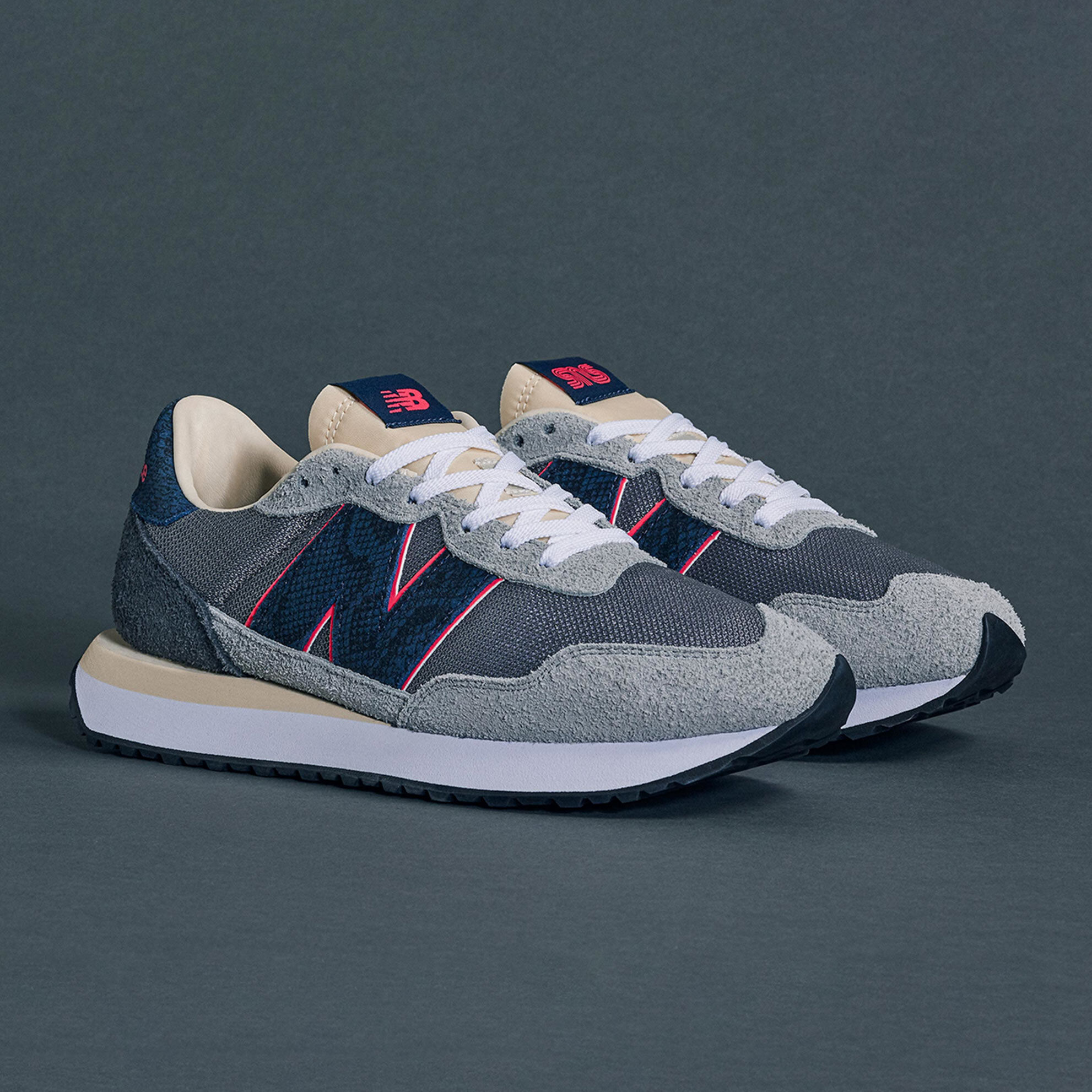 Une paire de chaussures de sport 'Blue Racer' 237 NB x SNS grises et bleu marine rehaussées d'accents magenta dans un studio sur une toile de fond bleue foncée