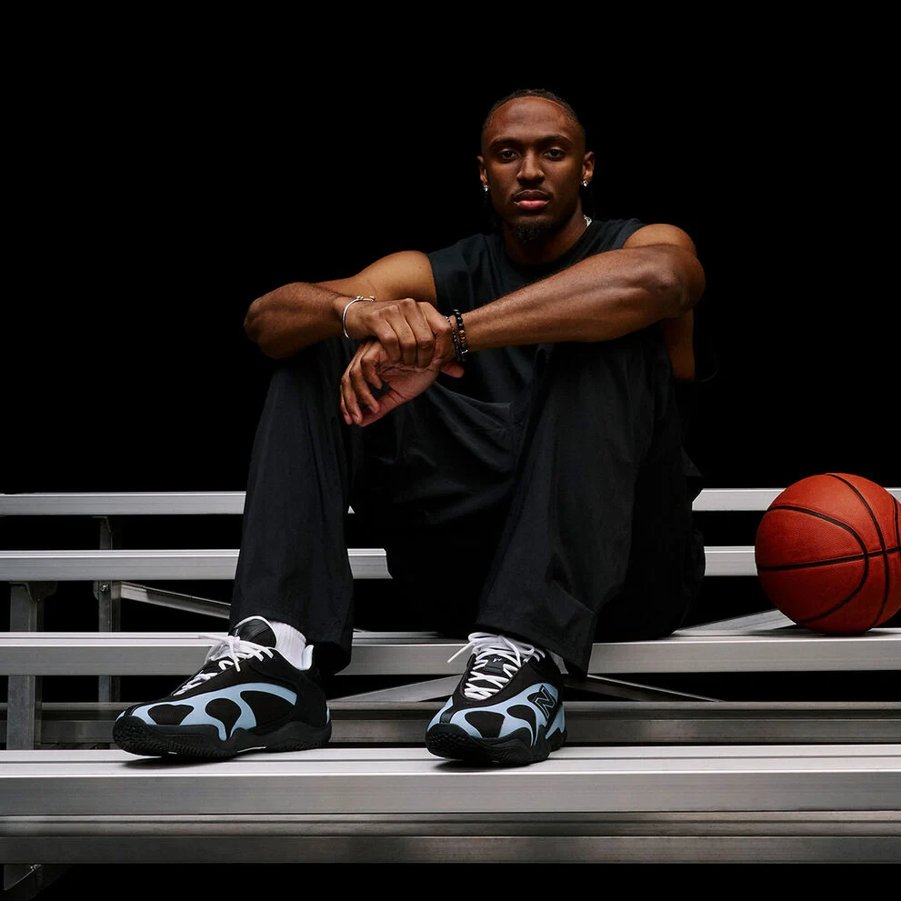 Person seated on metal bleachers beside a basketball, highlighting black-and-light-blue athletic sneakers in a dark studio setting.