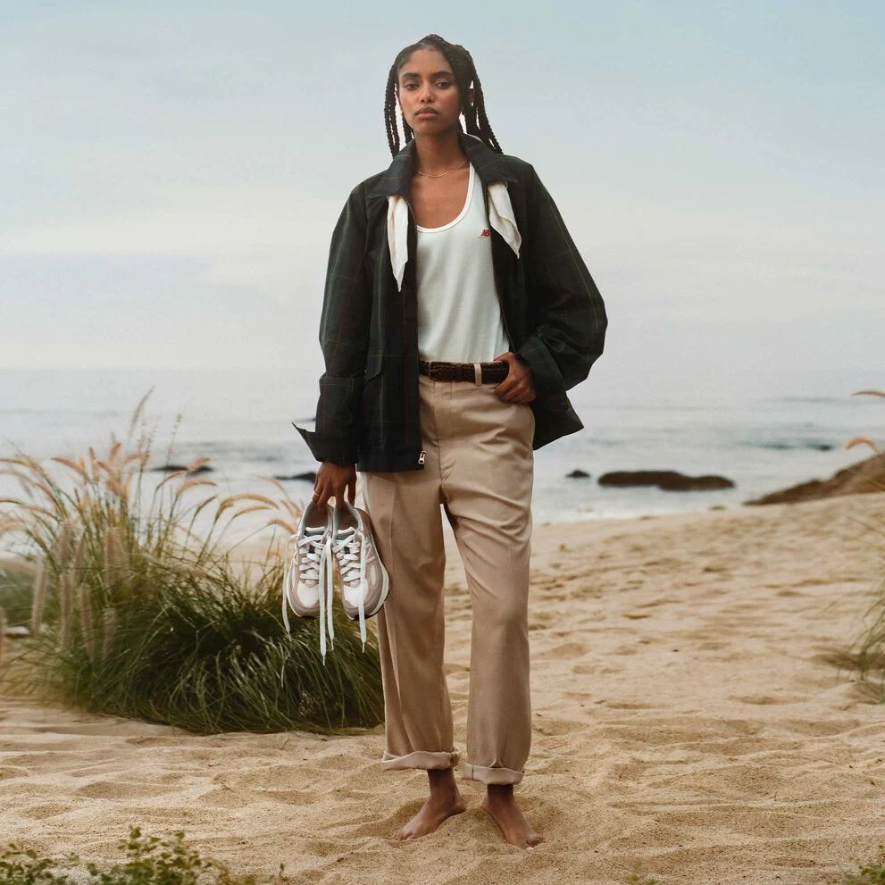 Une femme debout sur une plage, tenant une paire de chaussures 990v6. 