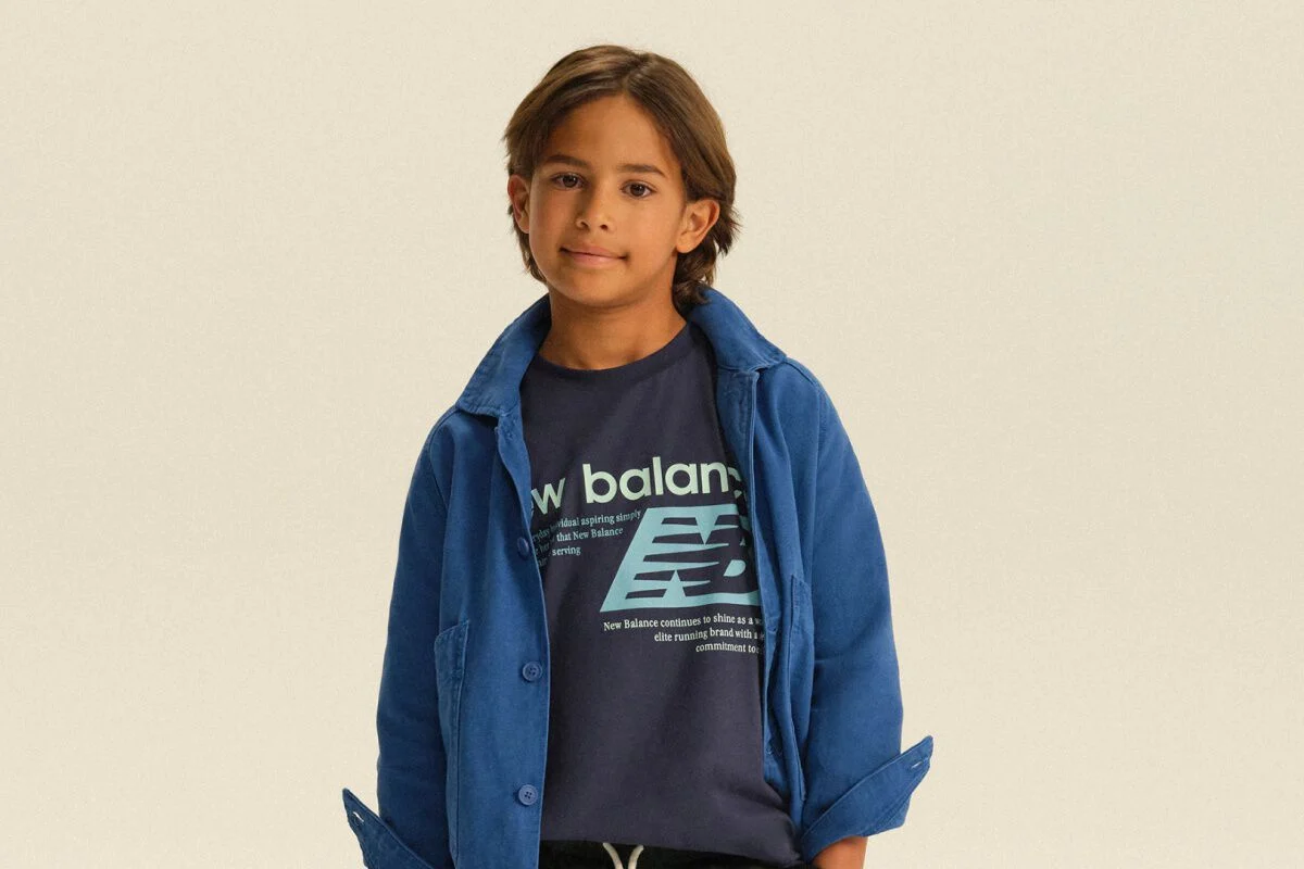 Enfant portant une surchemise bleue par-dessus un t-shirt New Balance &agrave; motif et un pantalon de jogging fonc&eacute;, dans un d&eacute;cor de studio &eacute;pur&eacute;.