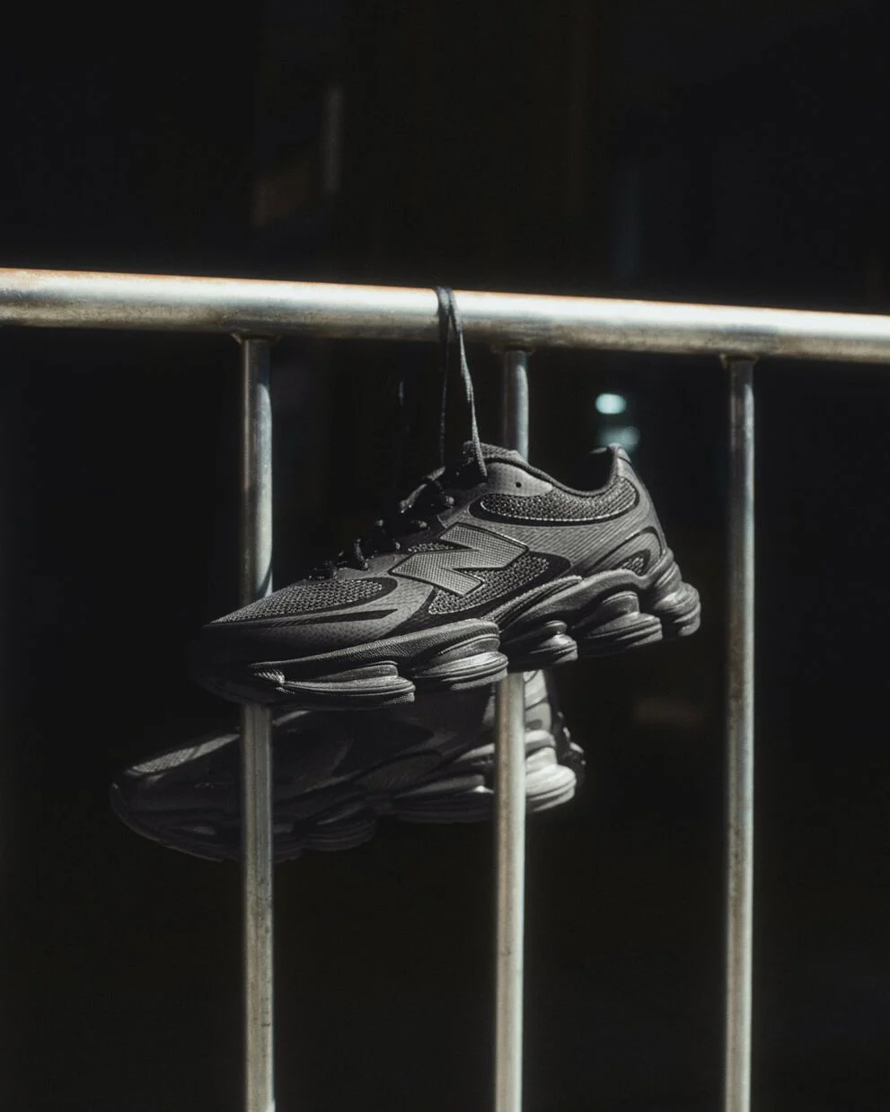 Black New Balance sneakers hanging by laces over a metal railing in dramatic lighting.