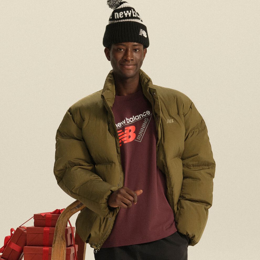 Person wearing an olive puffer jacket, burgundy New Balance shirt, and black knit beanie standing beside a wooden sled stacked with red gift boxes. 