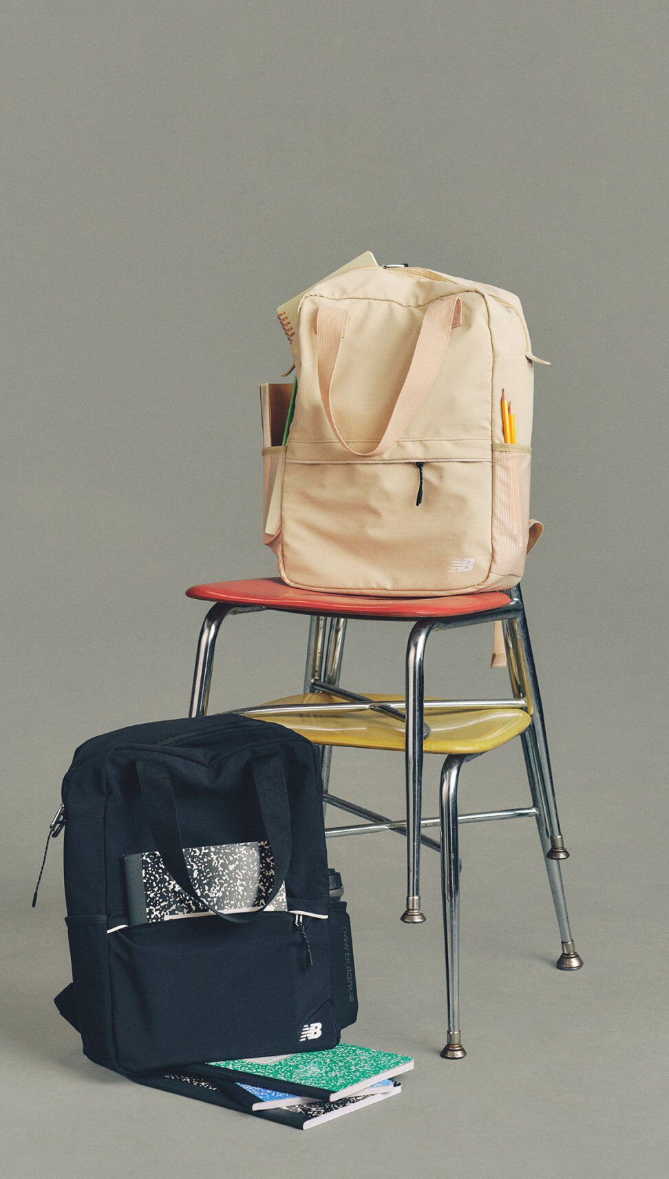 Studio image of two New Balance backpacks&mdash;one beige on stacked chairs and one black on the floor&mdash;surrounded by notebooks and school supplies against a neutral gray background.