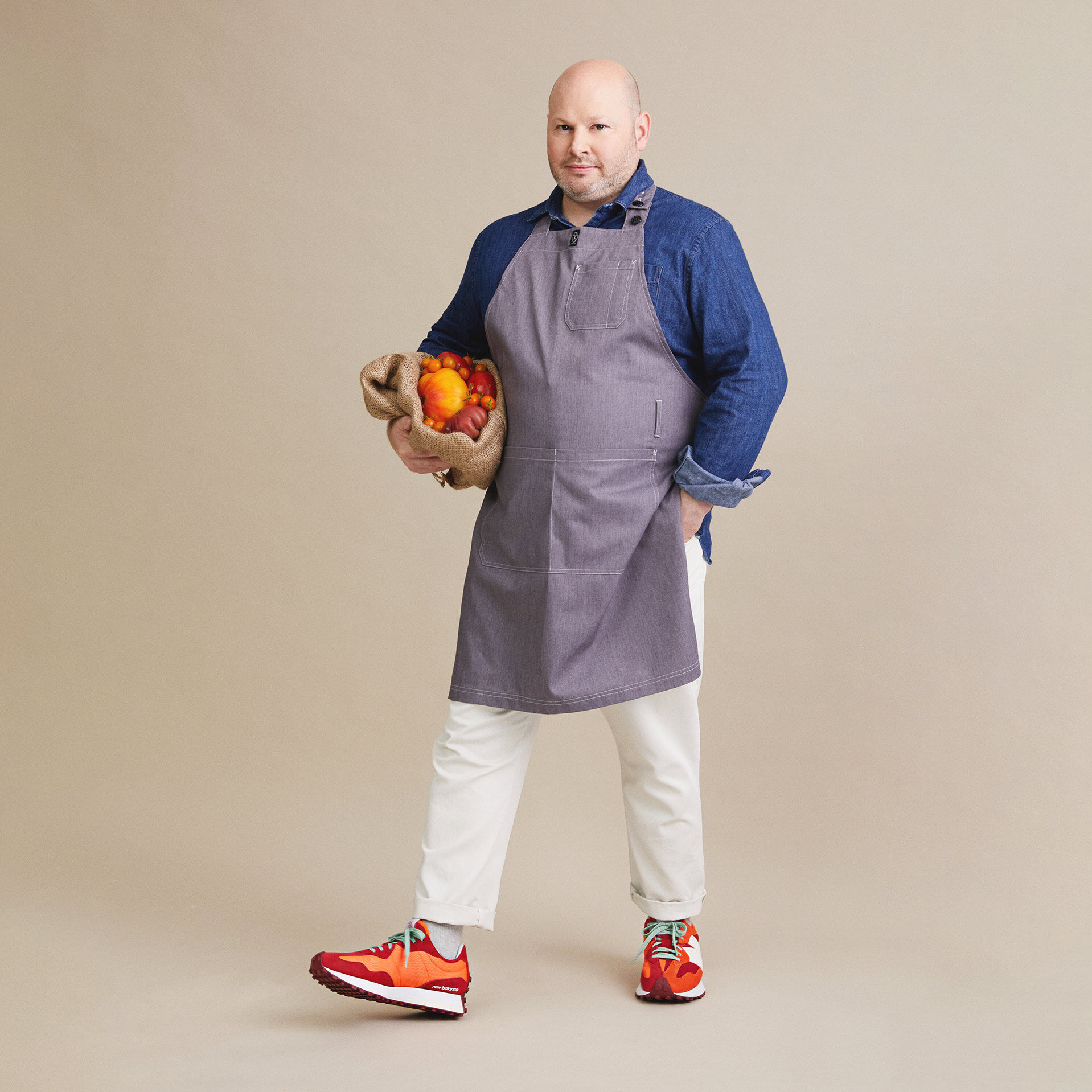 Debout dans un studio et vêtu de chaussures 327 oranges et rouges, le chef Dan Kluger tient à la main un sac en jute plein de fruits et légumes oranges et rouges