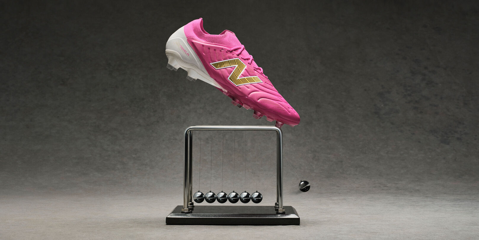 Bright pink New Balance soccer cleat with a metallic gold &ldquo;N&rdquo; logo appearing suspended above a black Newton&rsquo;s cradle on a gray studio surface, with one metal ball displaced to the side against a dark textured background.