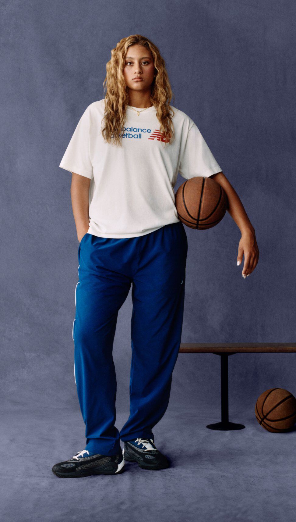 Une personne adopte une posture d&eacute;tendue devant un fond sombre textur&eacute;, portant un t-shirt de basketball New Balance blanc et un pantalon de sport bleu.