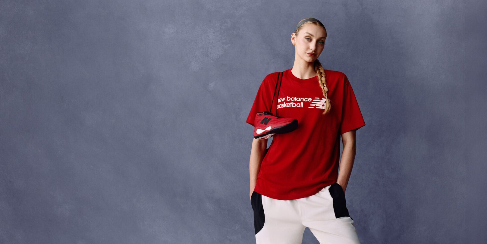 Athlete wearing a red New Balance basketball T-shirt holding a matching red and black sneaker against a gray studio background, with the &lsquo;new balance basketball&rsquo; logo visible on the shirt.