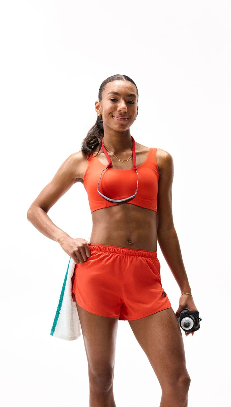 Person running in an orange color‑blocked bra and teal shorts, mid-stride against a clean white background. 