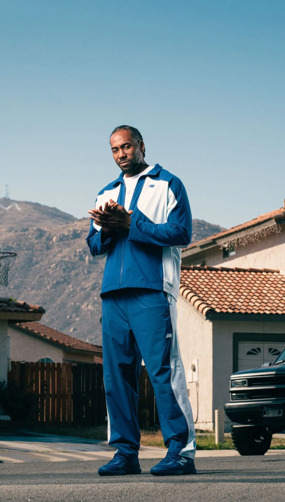 Une personne v&ecirc;tue d&rsquo;un ensemble de surv&ecirc;tement bleu et blanc et de chaussures de sport assorties se tient dans une rue de banlieue, avec des maisons et des montagnes en arri&egrave;re-plan.&nbsp;