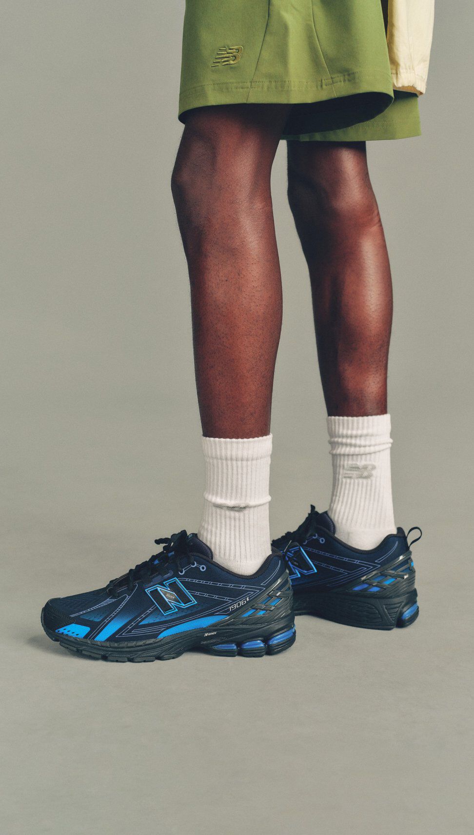 Close-up of a person standing in white crew socks and black-and-blue New Balance sneakers with visible cushioning, against a neutral background.