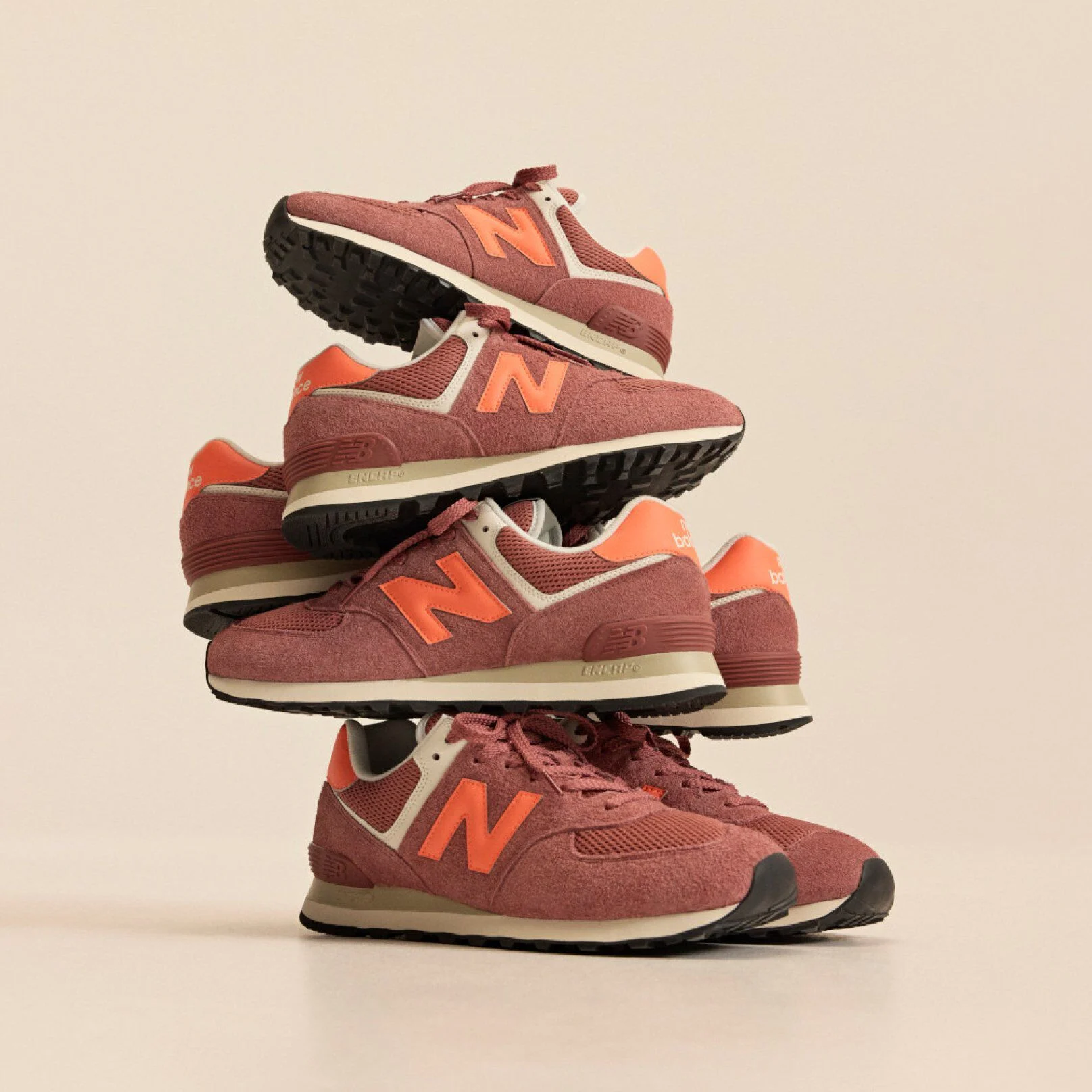 Stack of brick‑red New Balance sneakers with orange accents arranged in a balanced tower against a beige background.