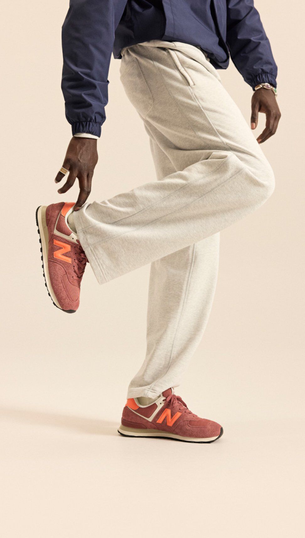 Person wearing brick‑red New Balance sneakers with orange accents, shown mid‑pose against a neutral background.