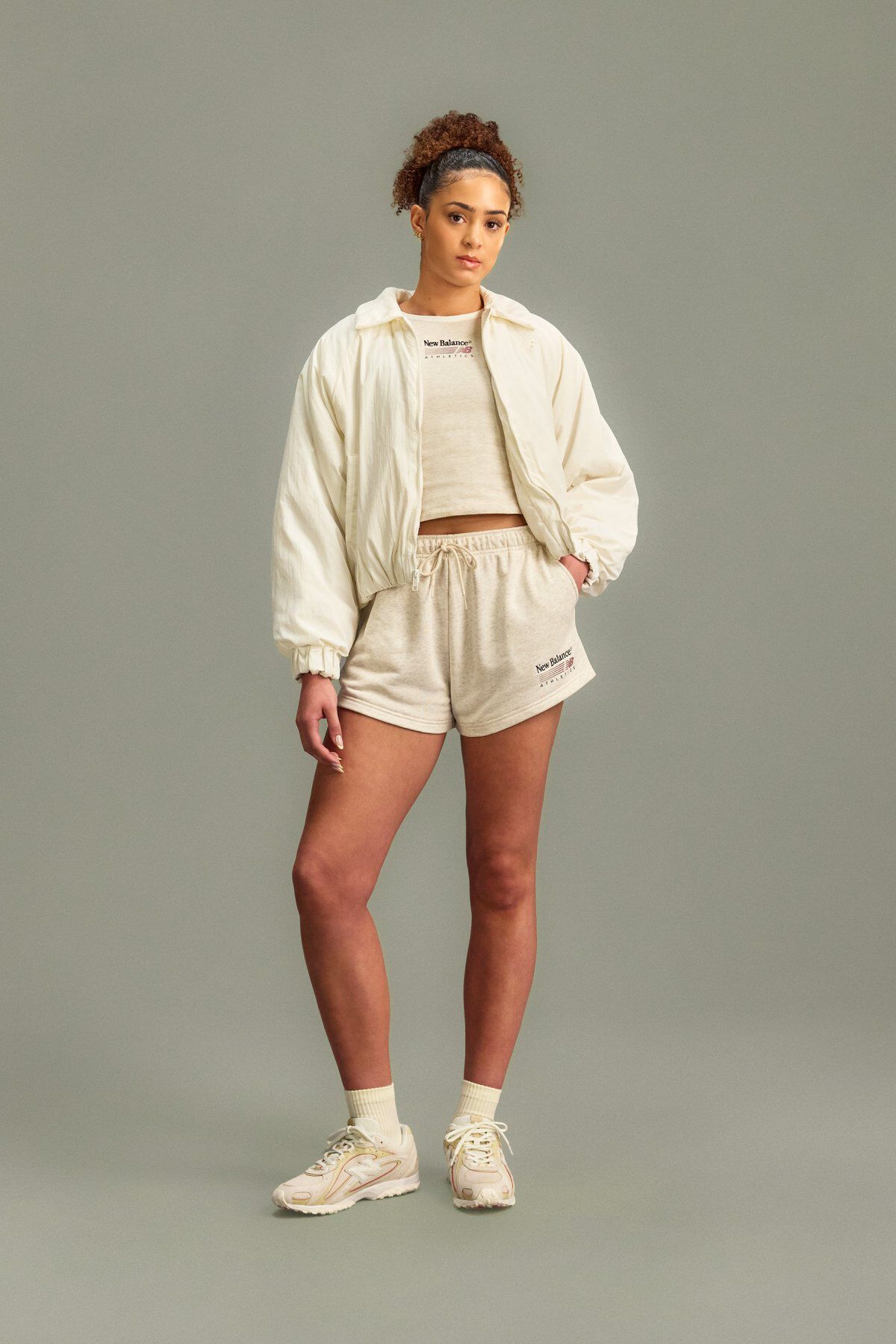 A woman wearing a New Balance shirt, jacket and shorts standing in front of a grey studio background.