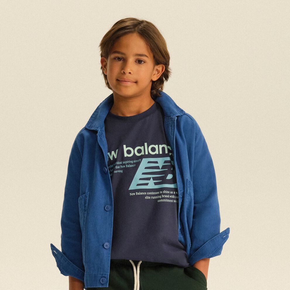 Enfant portant une surchemise bleue par-dessus un t-shirt New Balance &agrave; motif et un pantalon de jogging fonc&eacute;, dans un d&eacute;cor de studio &eacute;pur&eacute;.