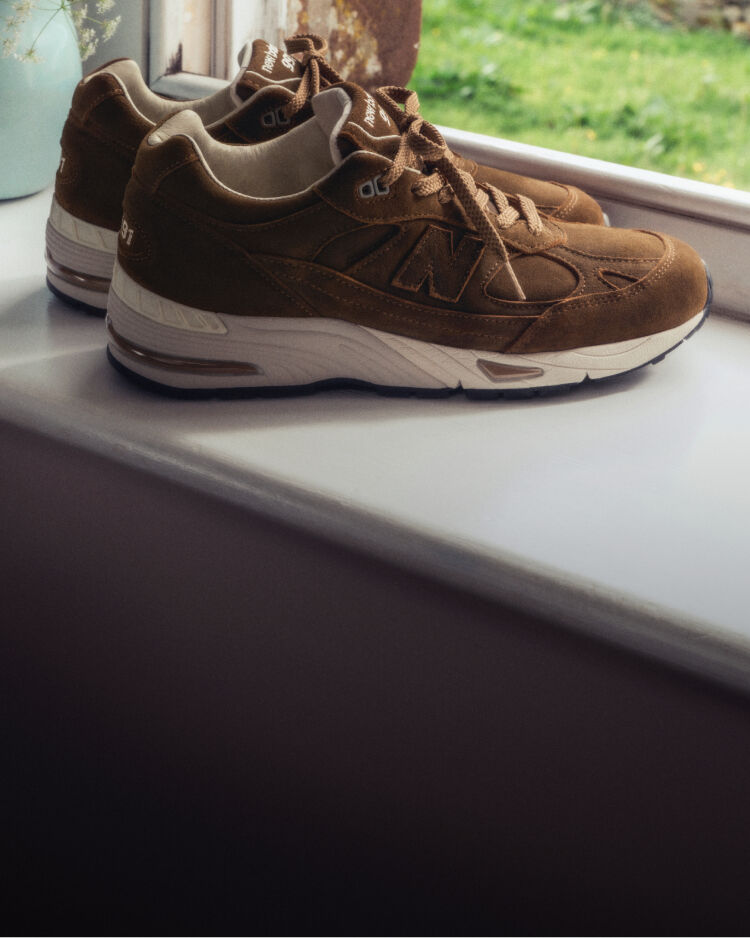 A pair of New Balance Made in UK sneakers on a windowsill next to a small vase of white flowers. 