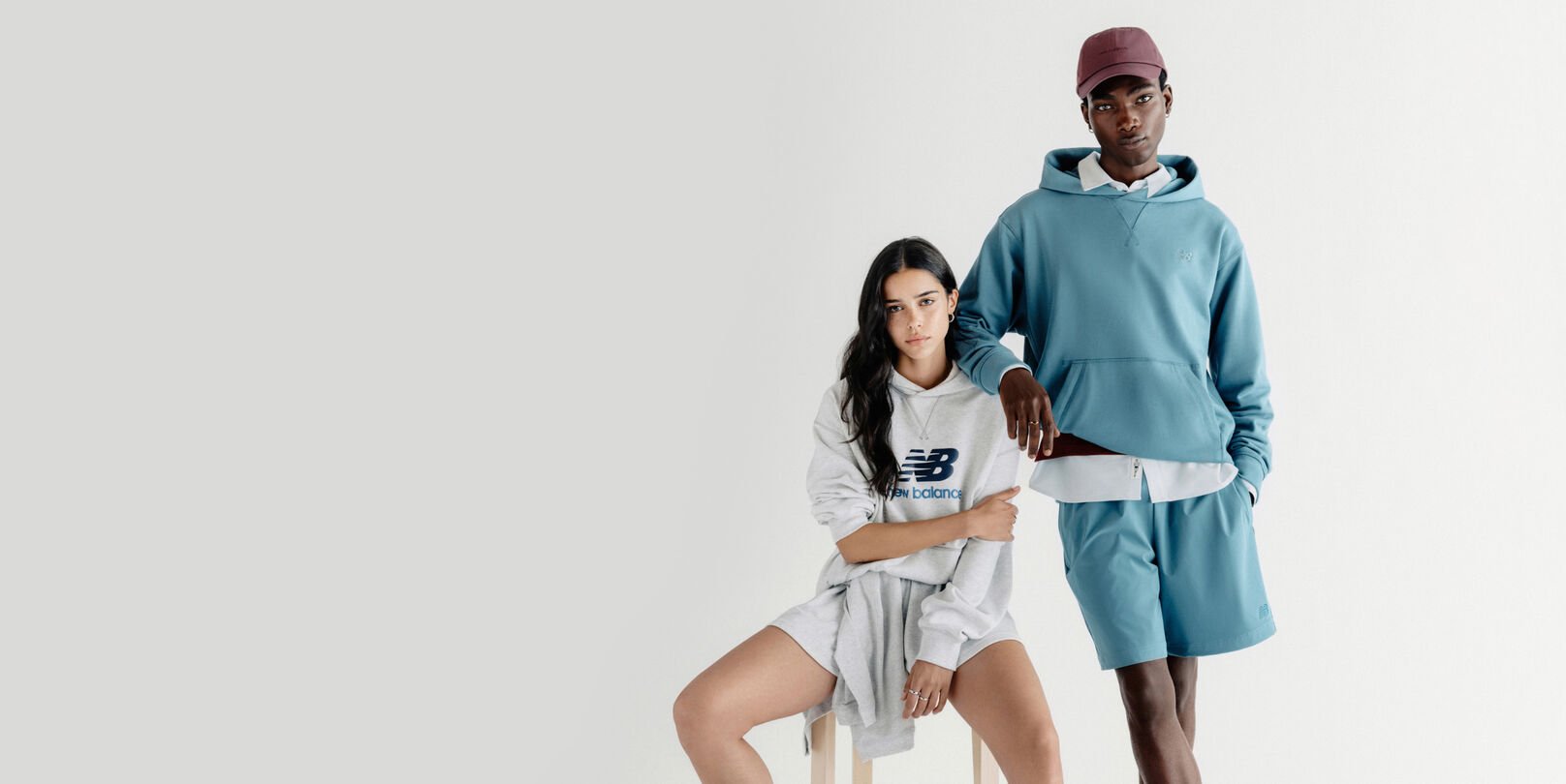 Un homme debout et une femme assise sur un tabouret devant un d&eacute;cor gris de studio.
