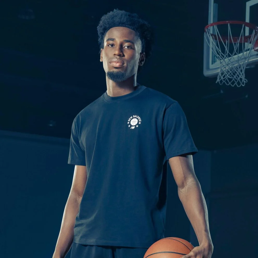 Une personne se tient sur un terrain de basket int&eacute;rieur, portant un t-shirt fonc&eacute; avec un petit logo circulaire &laquo;&nbsp;New Balance Basketball&nbsp;&raquo; sur la poitrine, tenant un ballon de basket sur le c&ocirc;t&eacute;.