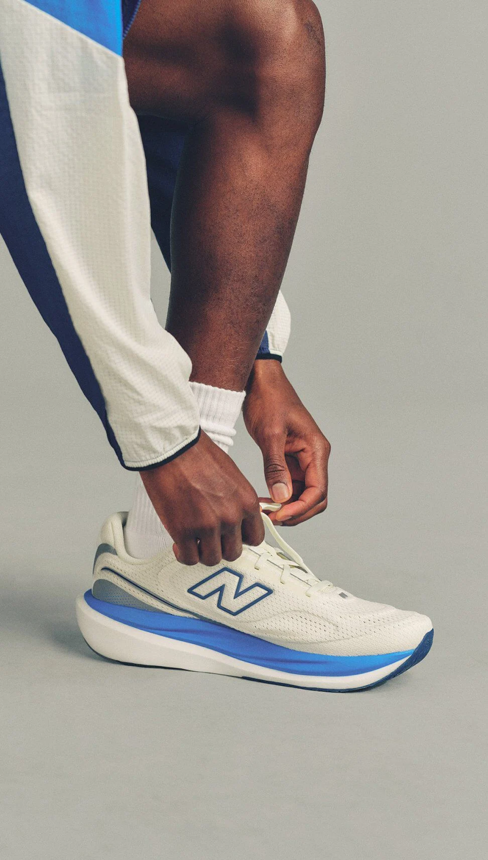 Close-up of a person tying white New Balance running shoes with blue accents while wearing white socks and light-colored athletic pants.