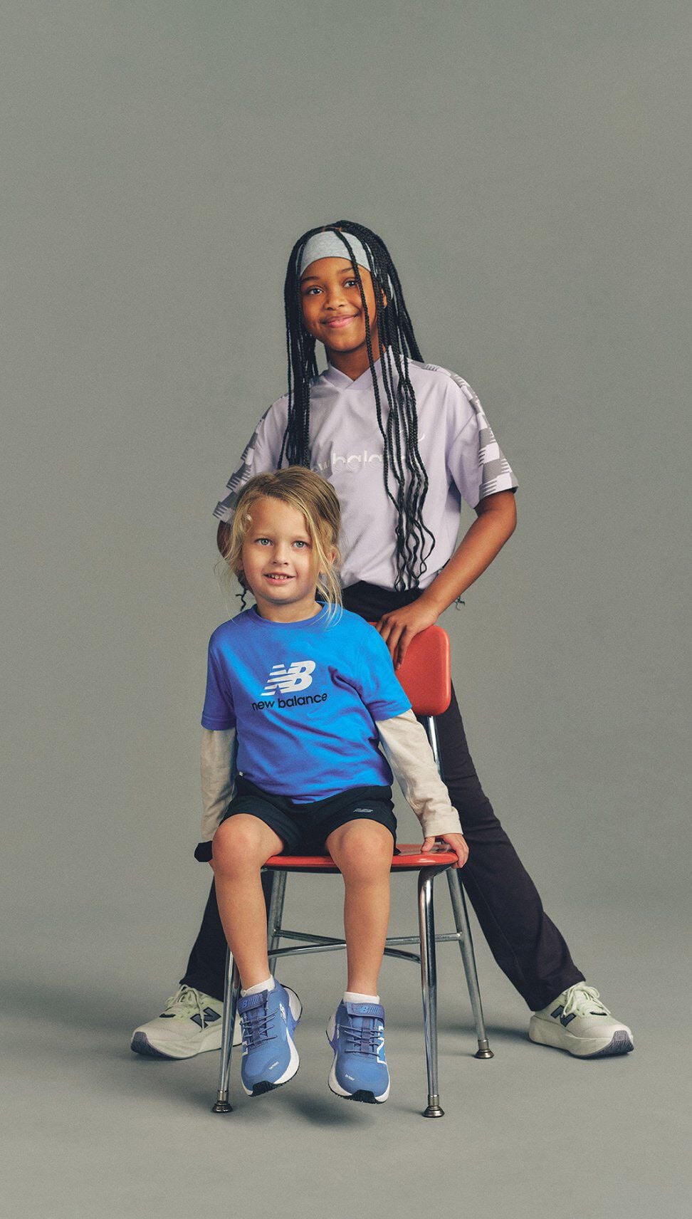 Two children in New Balance outfits pose with one seated on a chair in blue sneakers and the other standing behind with a hand on the chair against a neutral gray background.