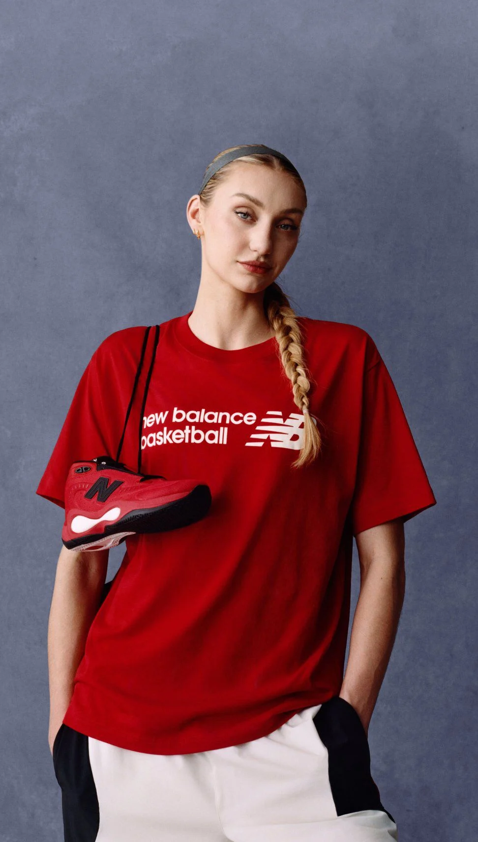 Un athl&egrave;te v&ecirc;tu d'un t-shirt de basket New Balance rouge tient une espadrille rouge et noire assortie devant un fond de studio gris ; le logo &laquo; New Balance Basketball&nbsp;&raquo; est visible sur le t-shirt.