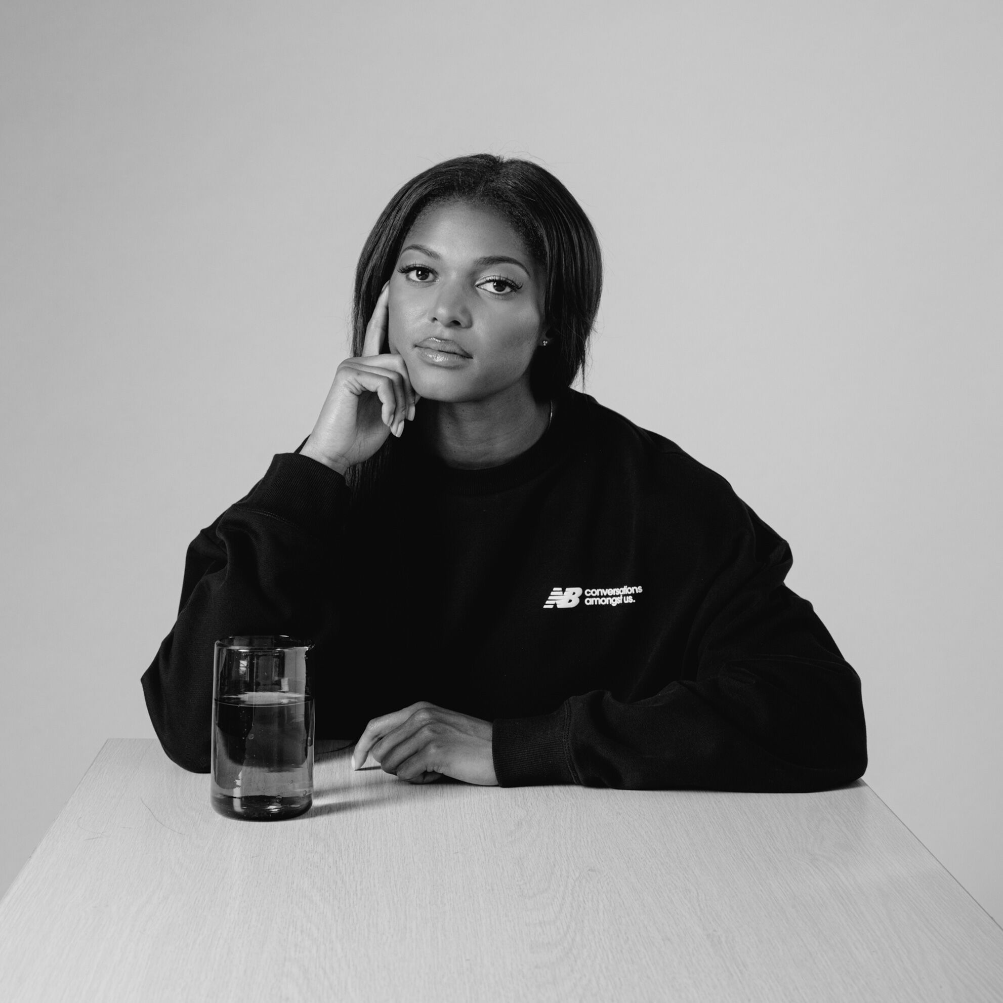 Black and white image of Gabby Thomas sitting at a table with a glass of water, wearing a black CAU hoodie