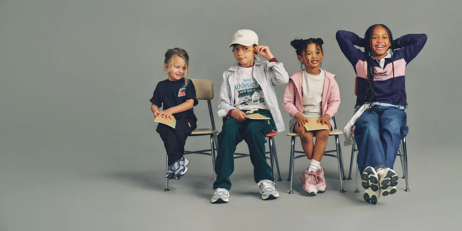 Four kids seated side-by-side on simple wooden chairs against a plain, neutral background, arranged in a horizontal line.
