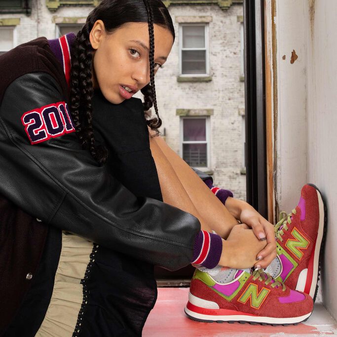 Assise sur un rebord de fenêtre, une jeune dame porte une veste de sport noire Stray Rats, une jupe ample noire et des chaussures de sport rouges NB x Stray Rats 574 aux accents roses et verts