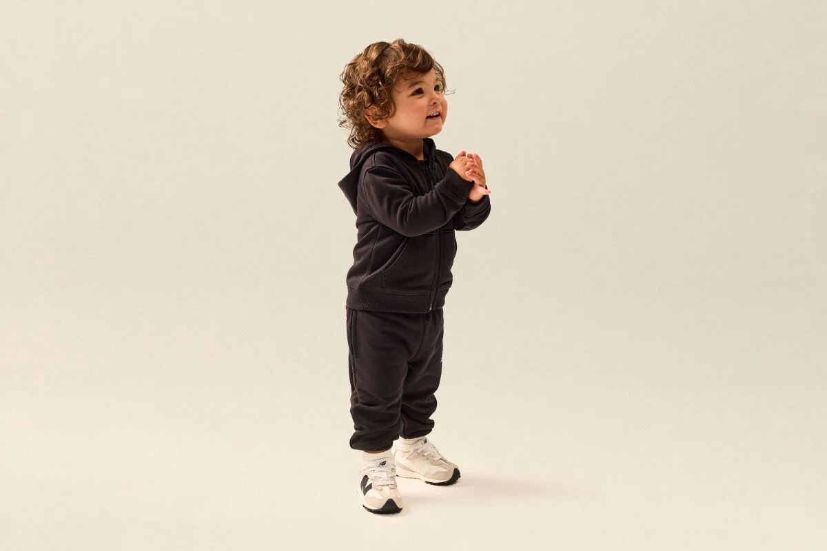 Tout-petit portant un ensemble molletonn&eacute; noir &agrave; capuchon et des chaussures de sport New Balance blanches, debout dans un d&eacute;cor de studio &eacute;pur&eacute;.