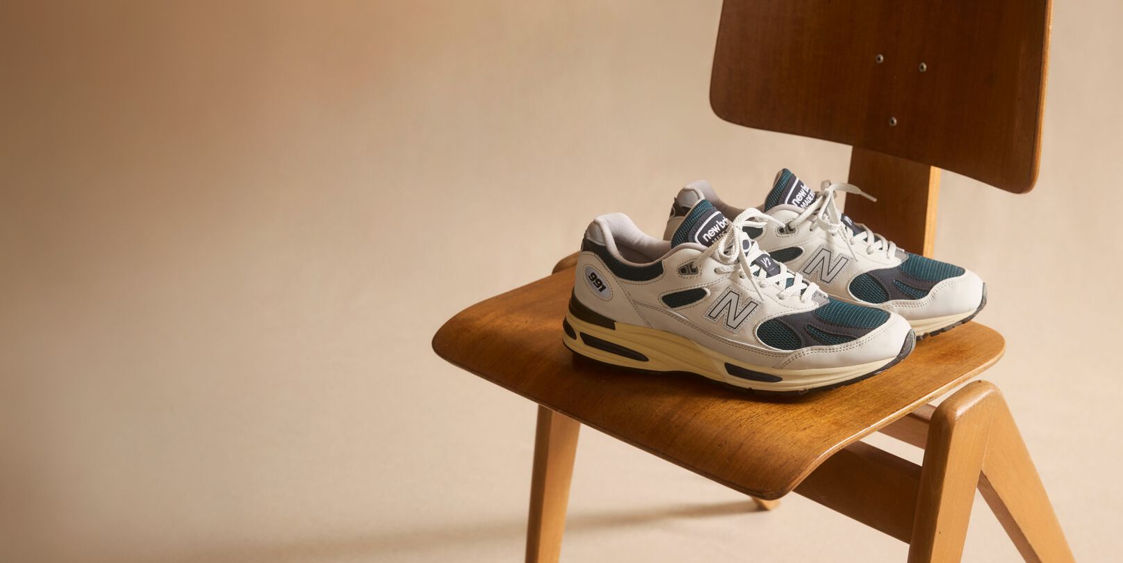 A pair of New Balance Made in UK sneakers sitting on a wooden chair in a beige studio environment. 