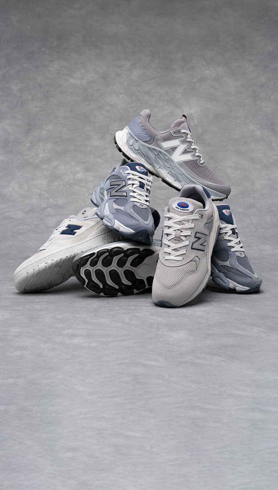 A group of grey New Balance sneakers stacked on each other in a grey studio environment.