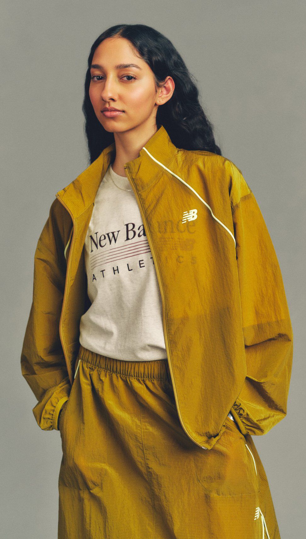 Close-up studio image of a person wearing a mustard-yellow New Balance windbreaker and matching pants over a graphic T-shirt against a neutral gray background.