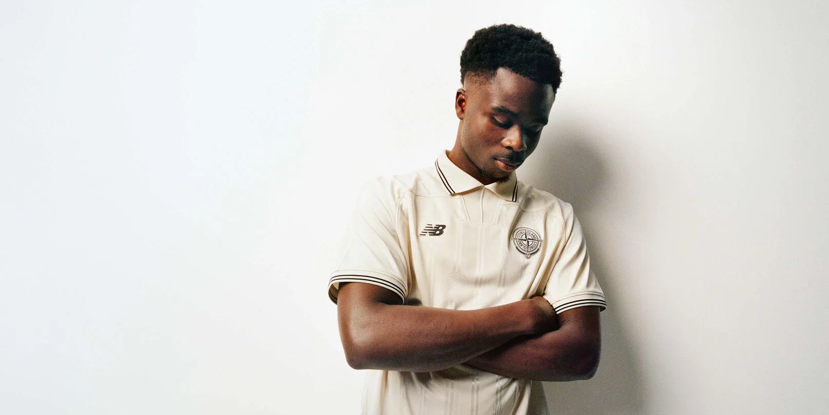 Saka looking down with his arms folded wearing a beige jersey.