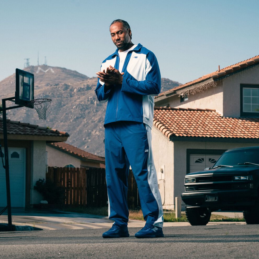 Une personne v&ecirc;tue d&rsquo;un ensemble de surv&ecirc;tement bleu et blanc et de chaussures de sport assorties se tient dans une rue de banlieue, avec des maisons et des montagnes en arri&egrave;re-plan.