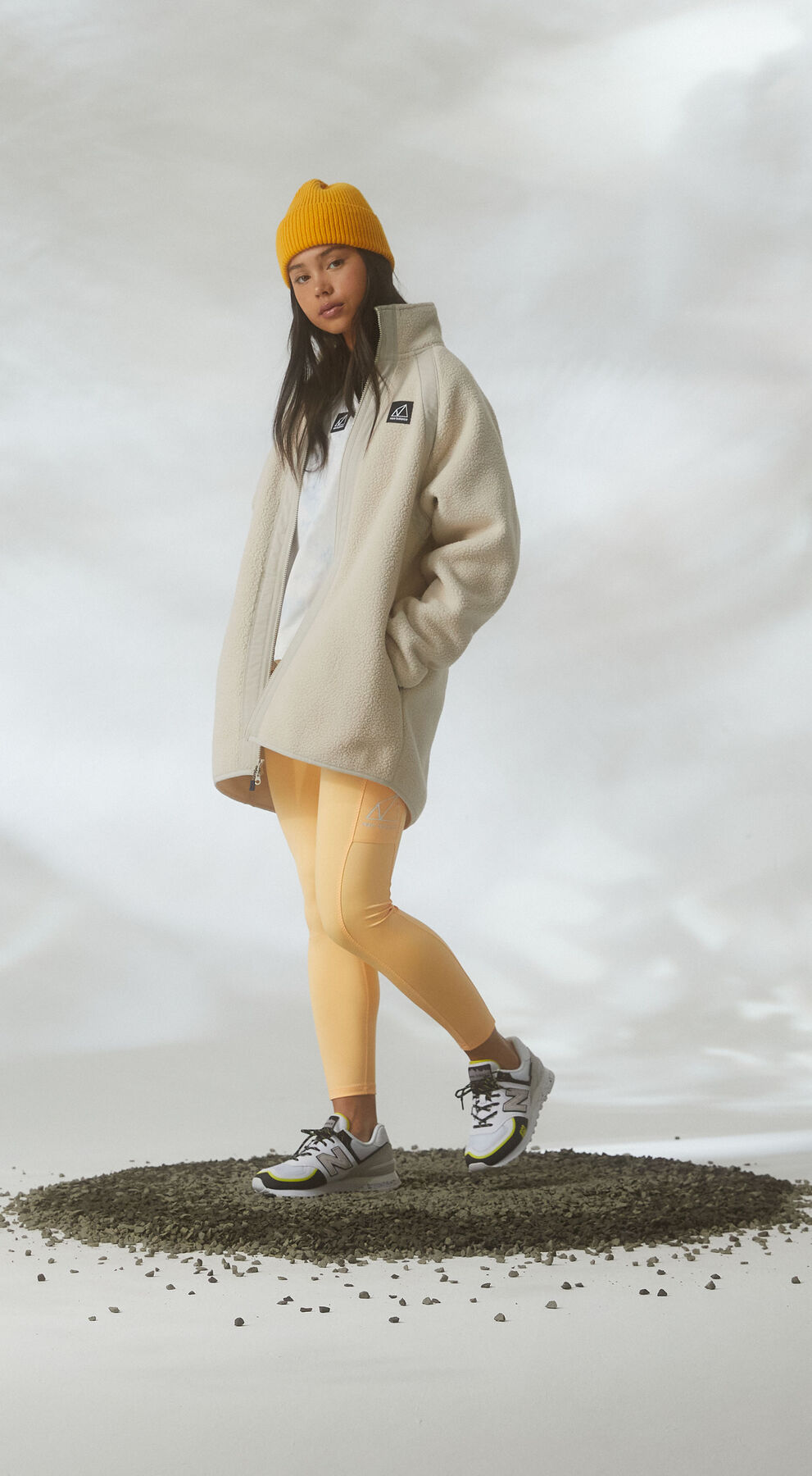 Une femme debout sur du gravier dans un décor de studio nuageux, portant des vêtements Tout-terrain et des chaussures 574.