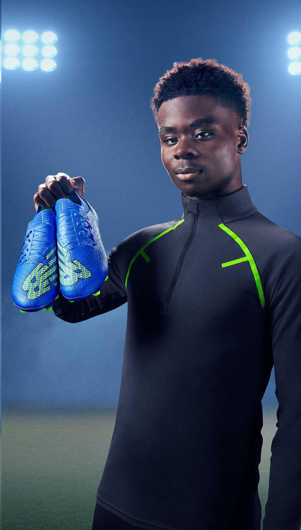 Raheem Sterling holding up a new pair of boots under lights on a field.