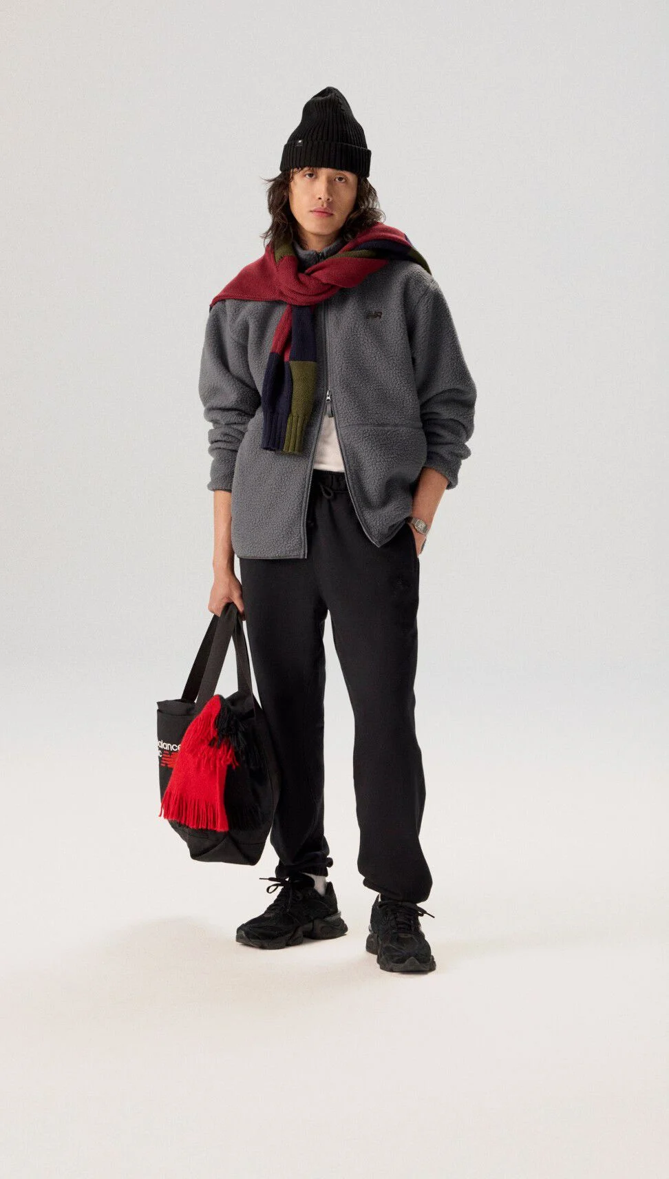 Jeune homme debout sur un fond gris, portant une veste grise tenant un sac.