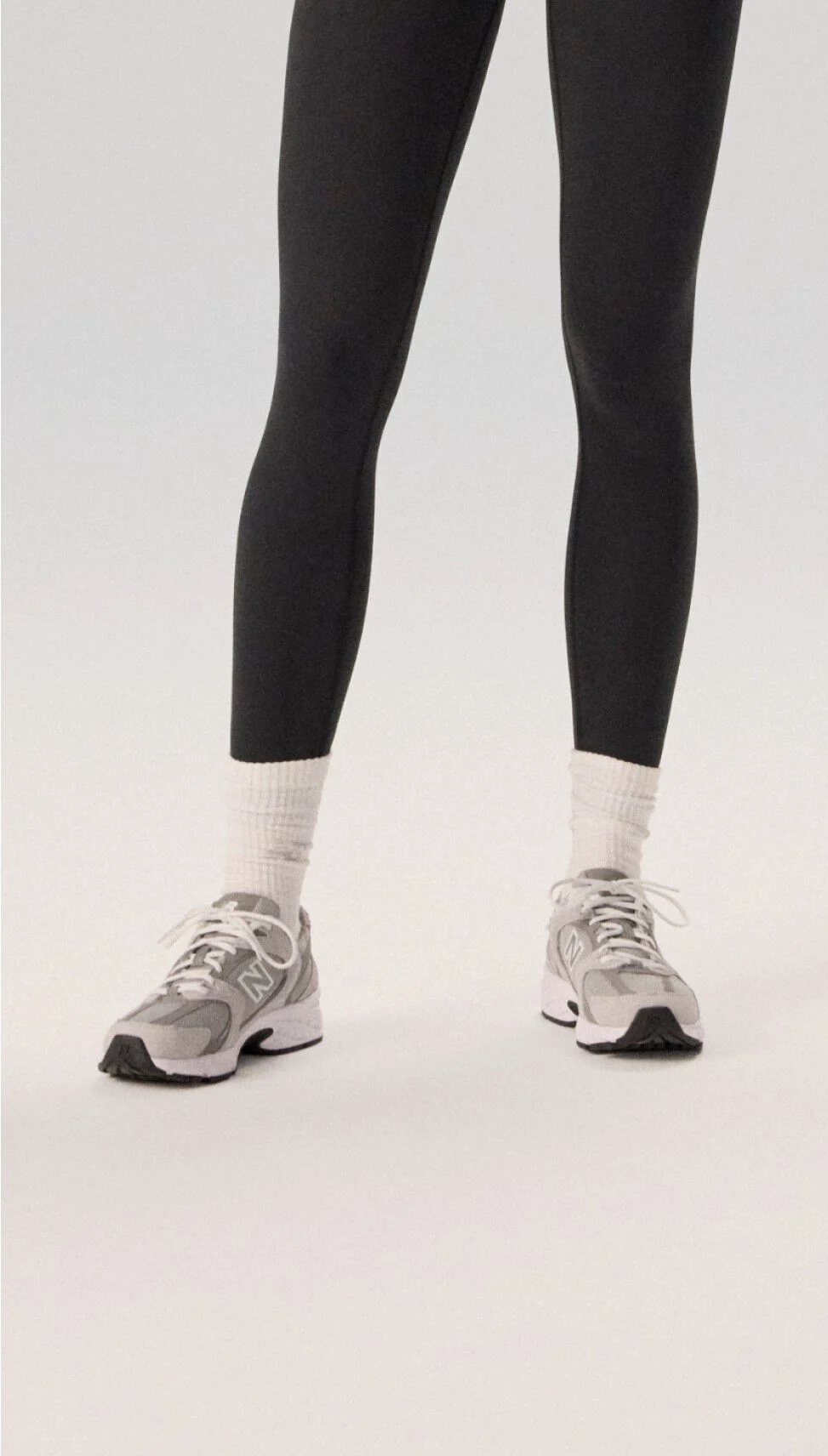 Femme portant des collants noirs et des chaussures de course grises sur un fond gris.