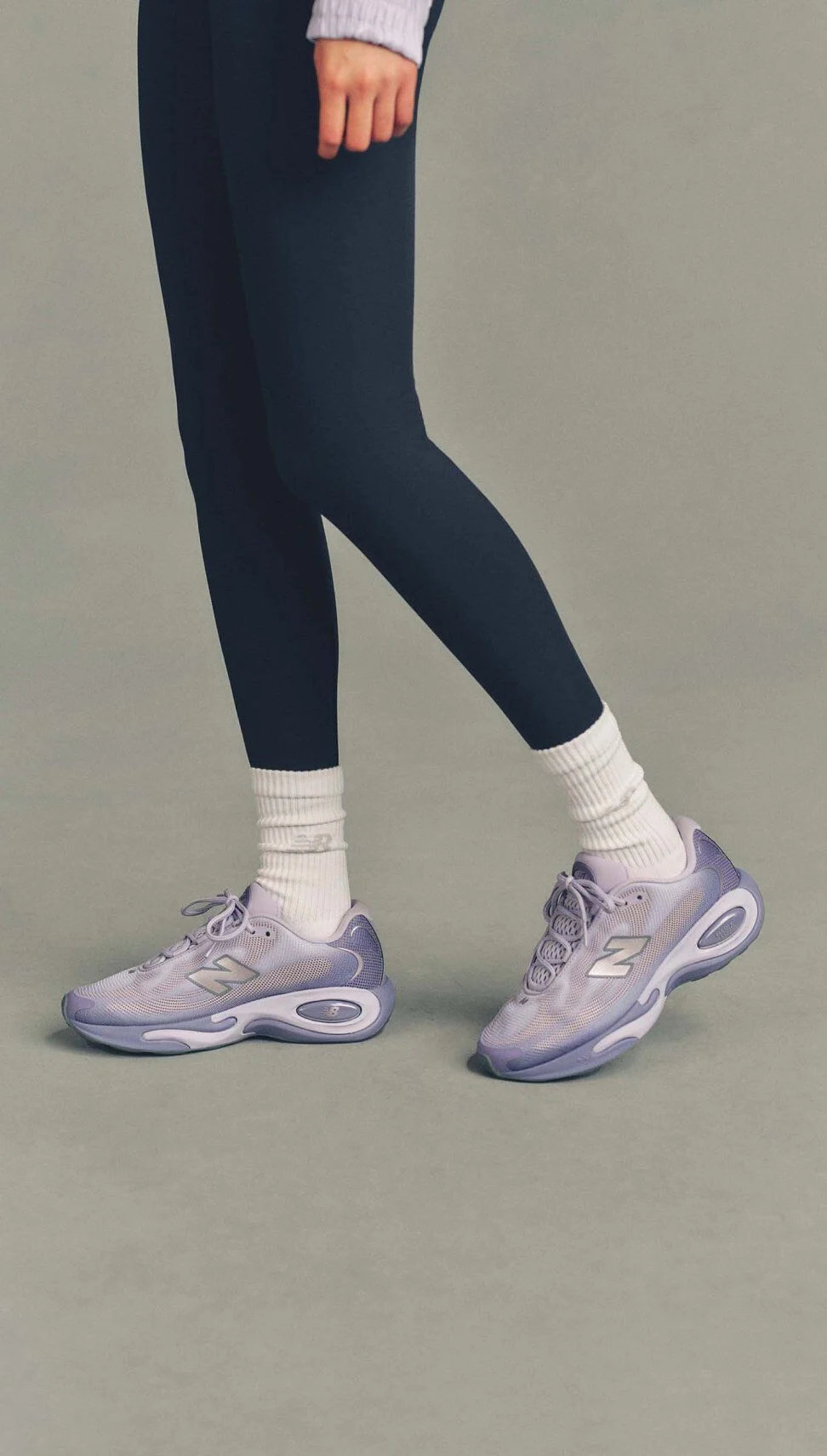 Close-up studio image of lavender New Balance running shoes paired with white socks and black leggings, with one foot slightly lifted against a neutral gray background.