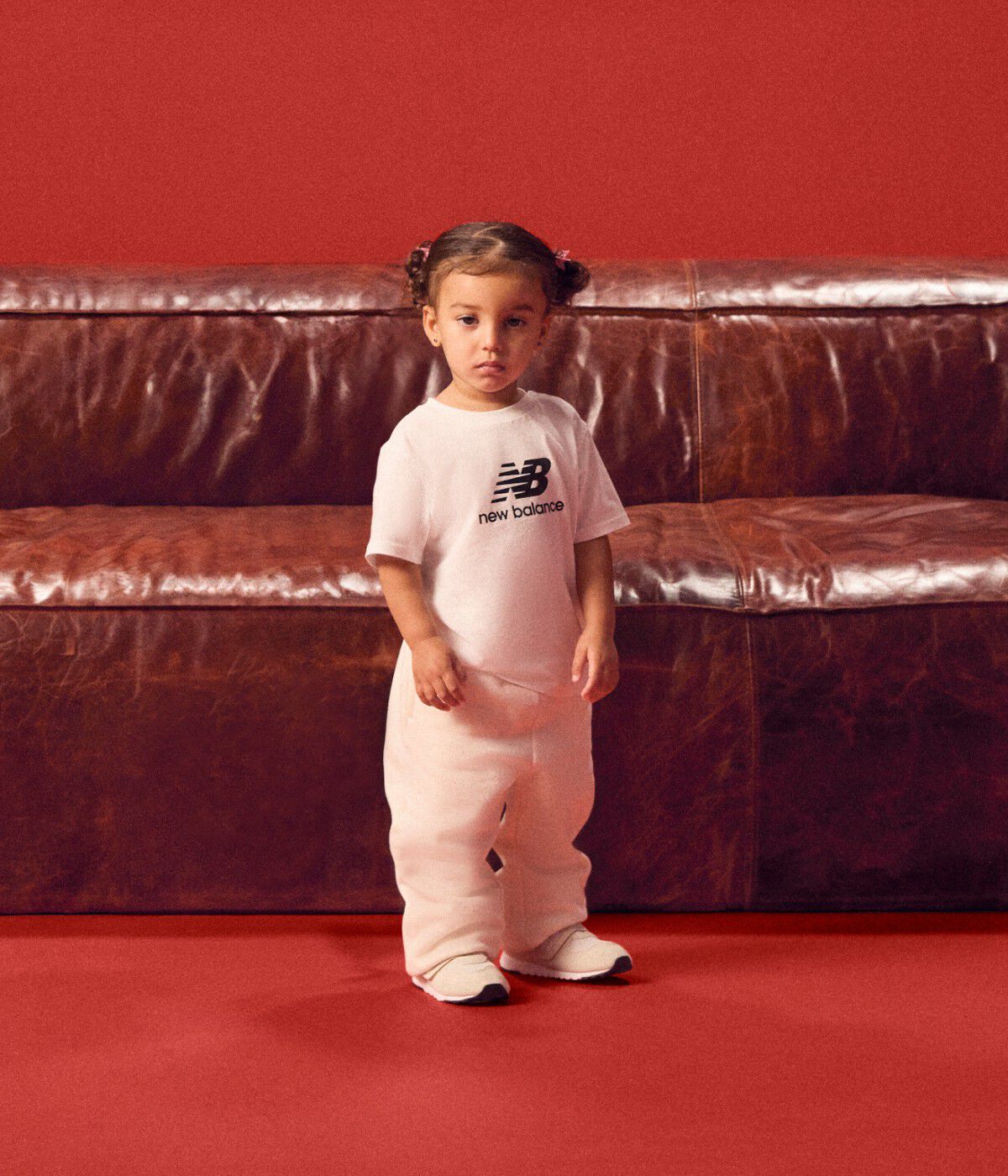 Baby standing in front of a red couch wearing a white tshirt.