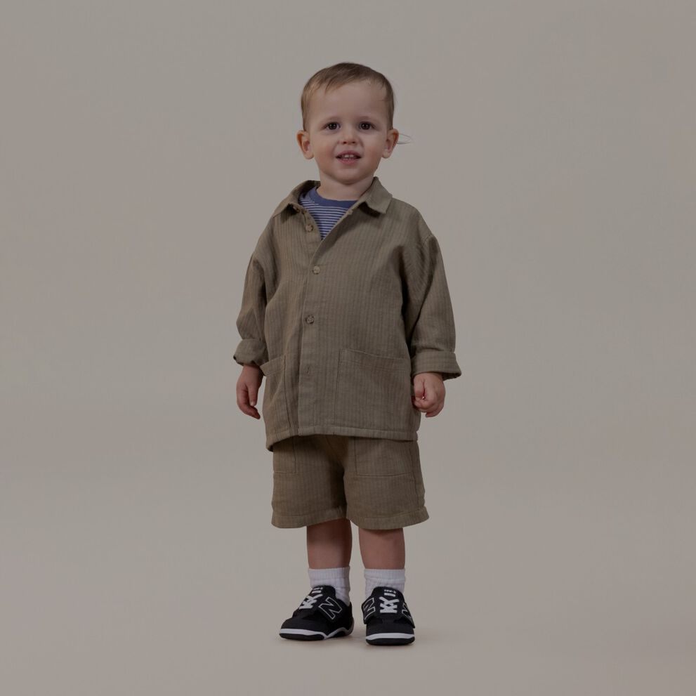 Little baby in a tan outfit standing on a grey background.