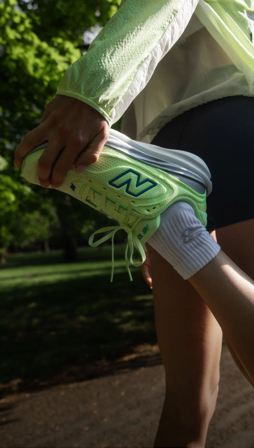 Close-up of a person stretching outdoors while holding a bright yellow-green New Balance running shoe with blue logo and white sole, wearing white socks and dark shorts. 