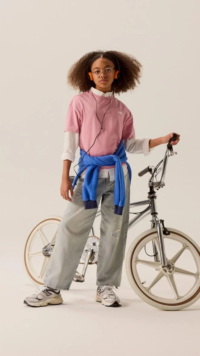 Young kid standing in front of a bike wearing blue jeans and a pink shirt. 