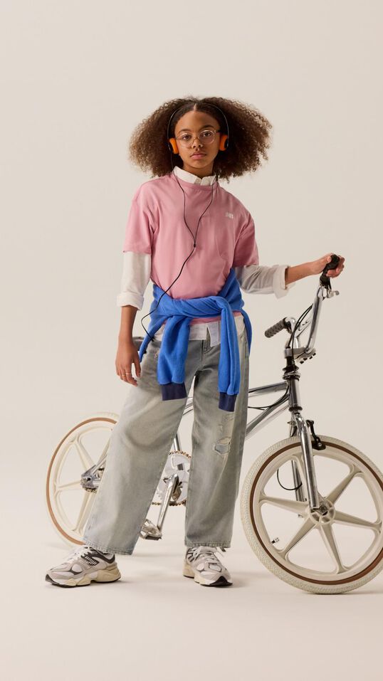 Jeune enfant debout devant un vélo portant un jean bleu et une chemise rose.