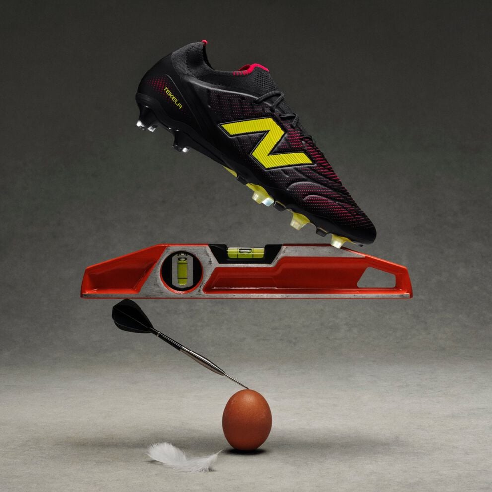 A black soccer cleat with yellow and red accents hovers above an orange leveling tool, which is balanced on a dart tip resting on a brown egg.