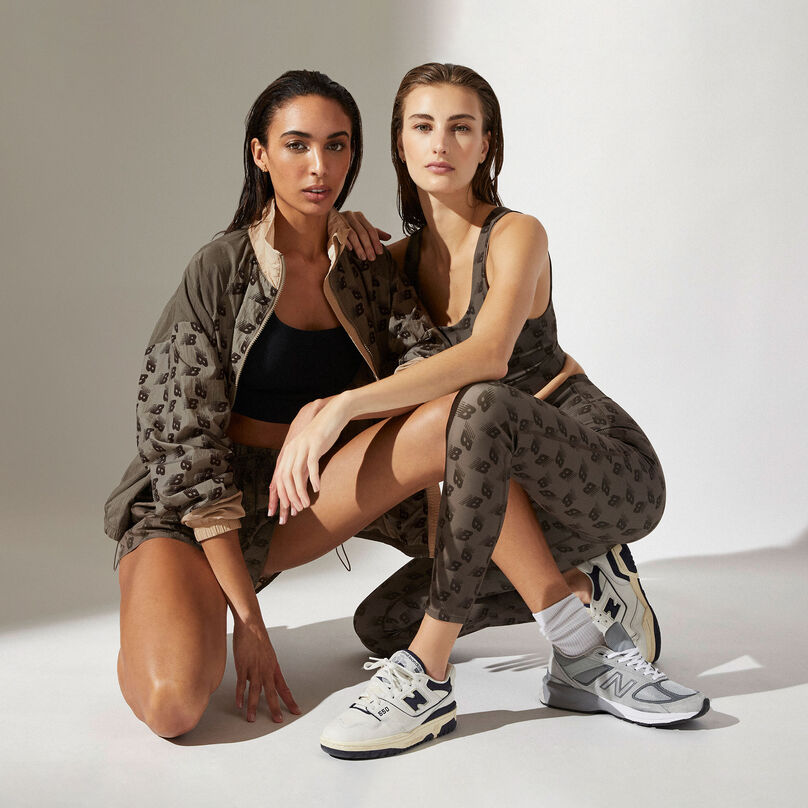 Two women wearing brown New Balance patterned clothing posed in squatting position against a grey studio background